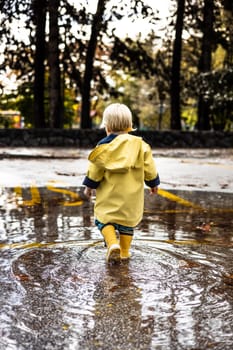 Small blond infant boy wearing yellow rubber boots and yellow waterproof raincoat walking in puddles on a overcast rainy day. Child in the rain