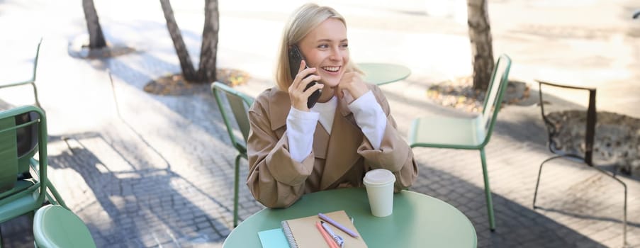 Carefree young woman in trench, talking on mobile phone and smiling, sitting outside cafe and drinking coffee, answer a call.