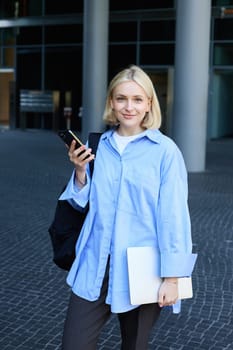 Vertical portrait of young office manager, woman in blue collar shirt and backpack, holding laptop and smartphone, waiting near business building on street, smiling at camera.