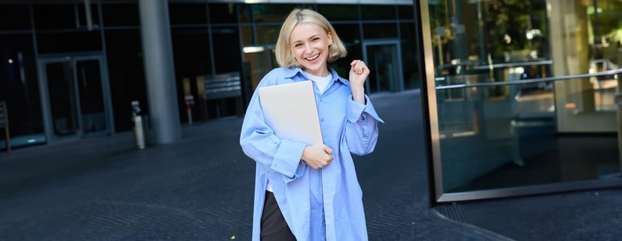 Portrait of young blond woman, student standing near her campus with notebooks and documents, wearing blue shirt and smiling at camera. Education concept