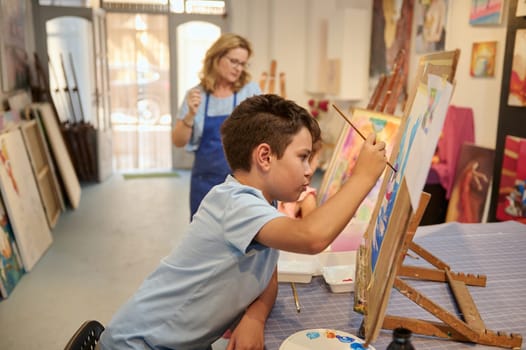 Authentic side portrait of a Caucasian handsome preteen school boy artist holding a paintbrush, focused on painting on easel in a creative art studio. Fine art. Drawing. Kids education concept