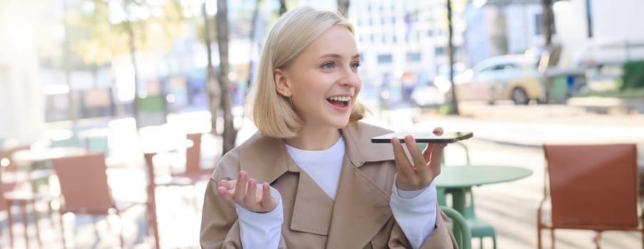 Lifestyle portrait of happy young woman, sitting in cafe and recording voice message, holding mobile phone near lips and talking into microphone, spending time in cafe, drinking coffee and chatting.