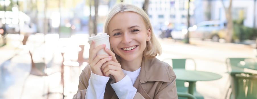 Close up portrait of smiling blonde girl, student sitting in outdoor cafe, holding cup of takeaway coffee near face, looking happy.