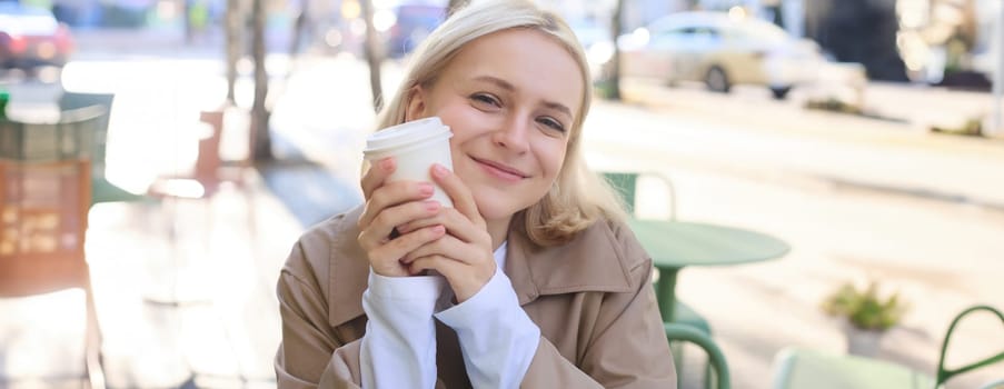 Close up portrait of smiling blond woman, holding cup of coffee, sitting in outdoor, street cafe, looking happy, waiting for someone in restaurant outside.