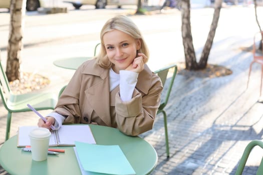 Modern student working on essay in outdoor cafe, doing homework, sitting alone and writing, drinking cup of coffee, smiling at camera.