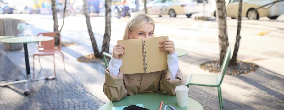 Portrait of young woman sitting in outdoor cafe, hiding behind journal, holding notebook in hands and smiling.