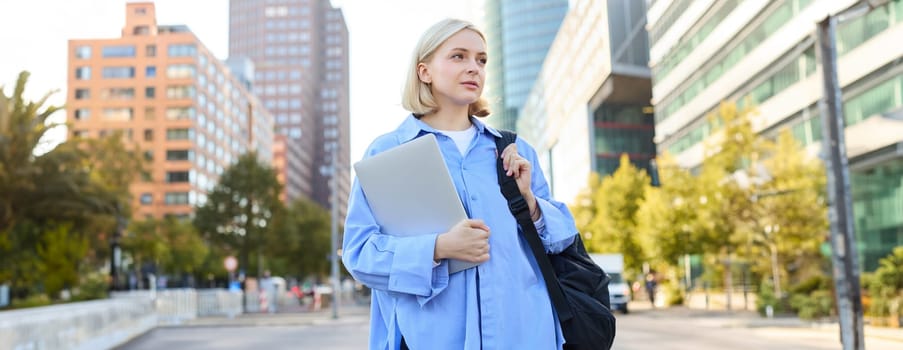 Portrait of smiling beautiful woman with backpack, holding laptop and looking around, walking along city streets, going to work on sunny warm day.