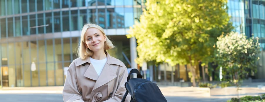 Vertical shot of young blond woman sitting on street bench with mobile phone, has backpack, waiting for someone, order taxi in smartphone application and looking around, smiling carefree.