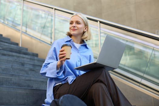 Lifestyle shot of young woman sitting on stairs with laptop, studying outdoors, drinking cup of coffee while working on computer, e-learning, studying on street.