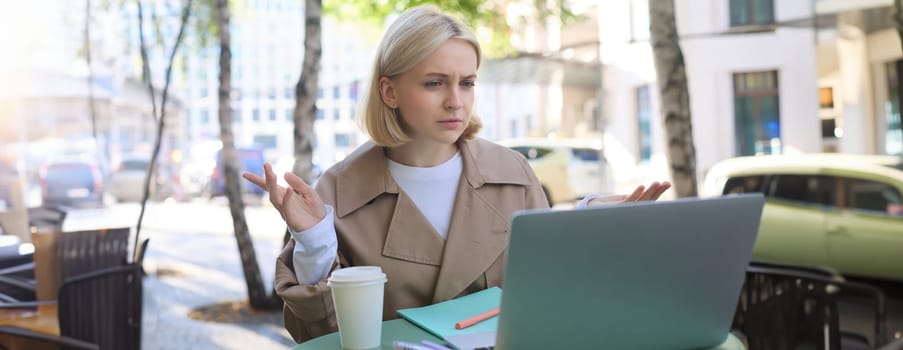 Image of woman with confused face, looking at laptop, working remotely, chatting with someone and shrugging, talking with puzzled face, sitting in coffee shop.