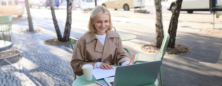 Happy, smiling young woman in trench, sitting in street cafe outdoors, drinking coffee, freelancing, looking at laptop and making notes, writing in notebook, working remotely.