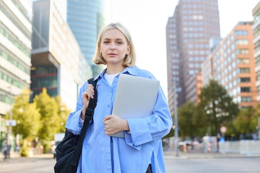 Portrait of young confident woman, college student with backpack and laptop, heading to lesson, standing outdoors on empty street with big buildings behind.