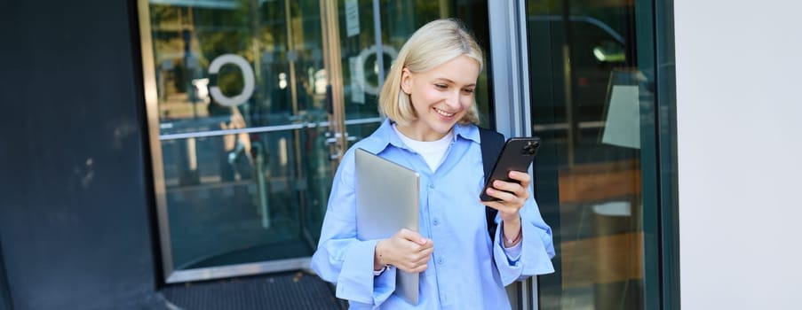 Image of young blond woman in blue shirt, holding laptop, waiting for someone near office building, using smartphone, mobile phone application.