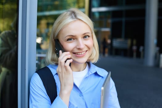 Close up portrait of modern young woman, student standing near building in city centre, talking on mobile phone, having chat on smartphone and smiling.