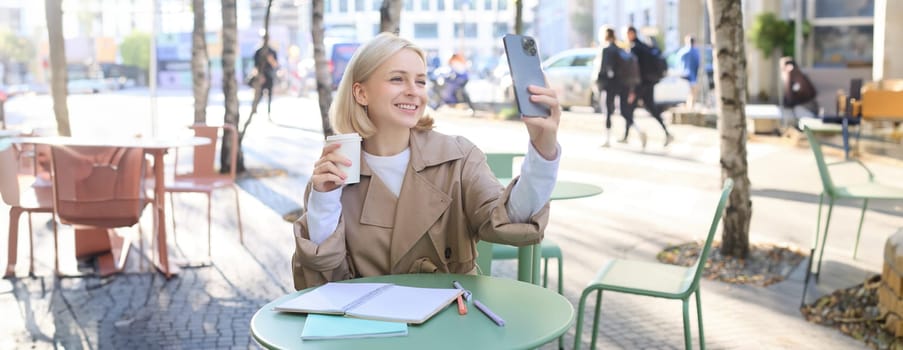 Happy blond woman taking selfie on smartphone, sitting outdoors in cafe and drinking coffee, making a post on social media to promote favourite shop.