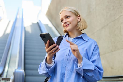 Close up portrait of young blond woman in city centre, standing near escalator, checking her mobile phone, reading message on smartphone. Lifestyle concept