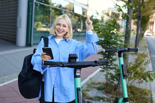 Portrait of young smiling female model, renting an electric scooter, using mobile phone to scan QR code with smartphone app, riding home from university, standing outdoors on street.