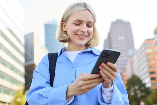 Close up portrait of stylish young blonde woman, standing on street, checking her mobile phone, using smartphone app to get around town, looking at online map for directions.