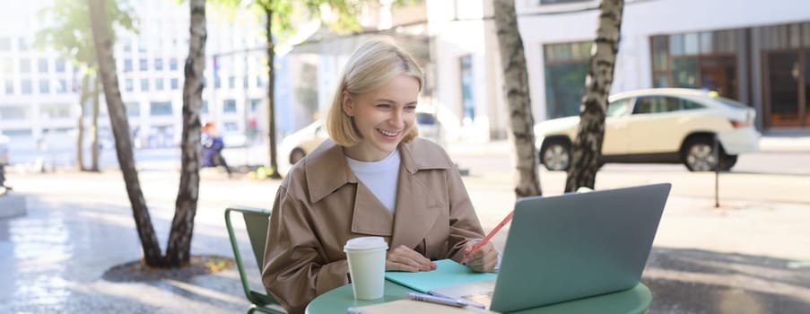 Young woman, freelancer, student doing homework in outdoor cafe, drinking her coffee on street, using laptop, connects to online lecture or doing course in internet.