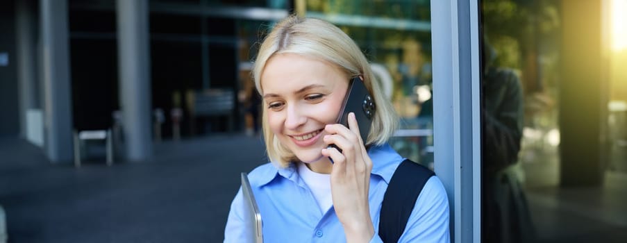 Happy female model, blond woman talking on mobile phone, chatting with someone, standing near office building with laptop and smartphone, smiling.