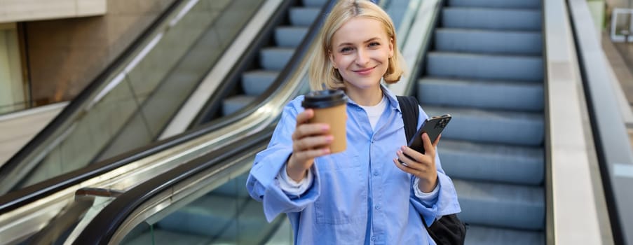 Portrait of smiling, beautiful young woman with smartphone, standing on bottom of escalator, giving you cup of coffee, brought takeaway.