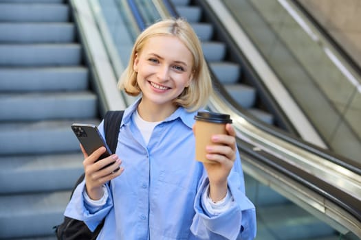 Portrait of young beautiful woman, girl student with cup of coffee and smartphone, standing near escalator in city centre, smiling and looking at camera.