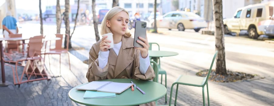 Image of stylish young woman, student taking selfie in cafe on street, posing with her favourite drink, enjoying coffee and making content for social media blog.