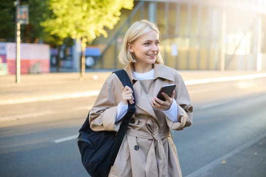 Modern young woman with backpack, student using smartphone, standing on street, sunny day, waiting for ride, using mobile phone application.