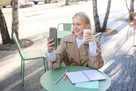 Image of stylish young woman, student taking selfie in cafe on street, posing with her favourite drink, enjoying coffee and making content for social media blog.