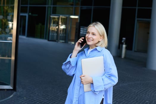 Image of young office woman, employee standing near company building with documents and notebooks, answers phone call, talking on smartphone and smiling, wearing blue collar shirt.