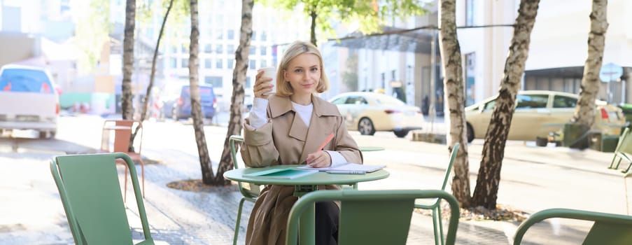Portrait of modern blond woman in trench coat, drinking coffee, holding takeaway cup, sitting in outdoor cafe and writing in notebook, looking serious at camera.