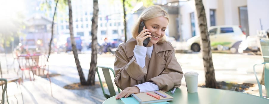 Portrait of young beautiful woman in cafe, sitting outdoors on bright sunny day, talking on smartphone, chatting over the phone and smiling.