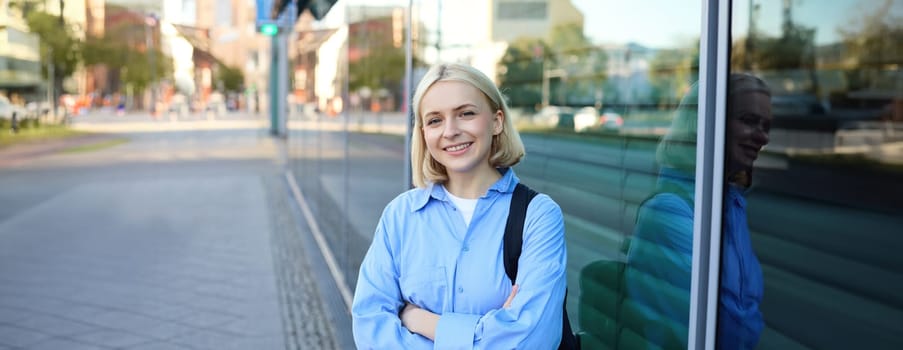 Lifestyle portrait of young smiling woman, student or office employee, standing on street in blue shirt, cross arms on chest and looking confident at camera.