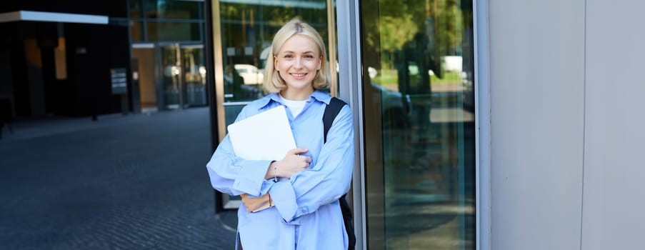 Image of young college girl, young woman with laptop, posing near university building, standing on street, smiling, concept of education and people.