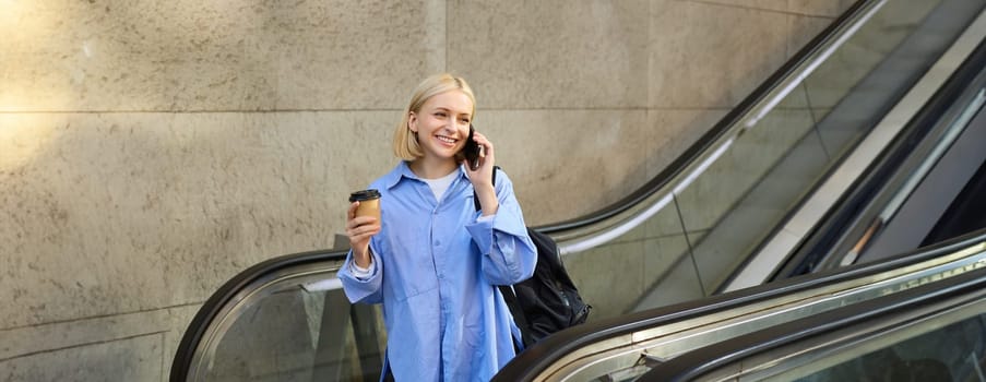 Street style shot of young beautiful woman, student on escalator going down to subway, talking on mobile phone and drinking coffee.