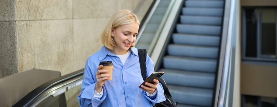 Portrait of blond young woman, reading message on her mobile phone, drinking takeaway coffee, standing near escalator with backpack.