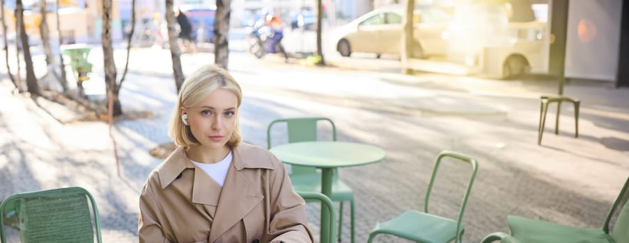 Vertical shot of young blond woman in wireless headphones, making notes, studying online, attend web lecture, using laptop, writing in notebook, looking serious at camera.