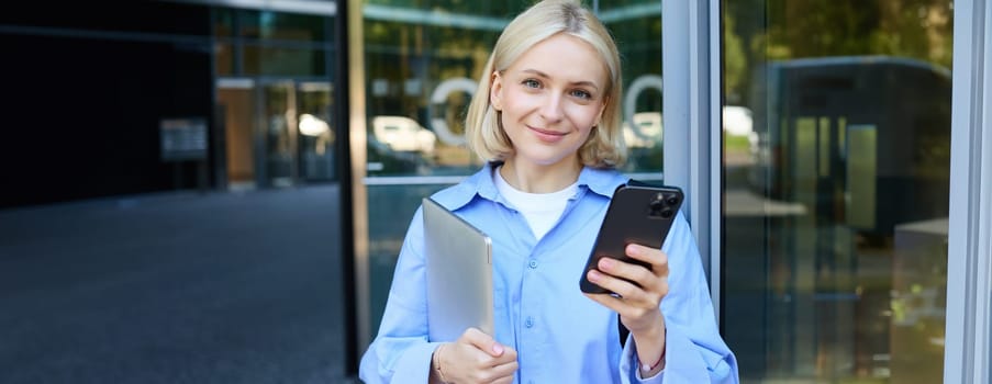 Portrait of young female student standing outside university campus, posing near building, holding laptop and smartphone, wearing blue collar shirt.