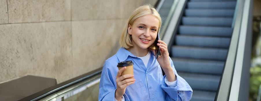 Street style shot of young beautiful woman, student on escalator going down to subway, talking on mobile phone and drinking coffee.