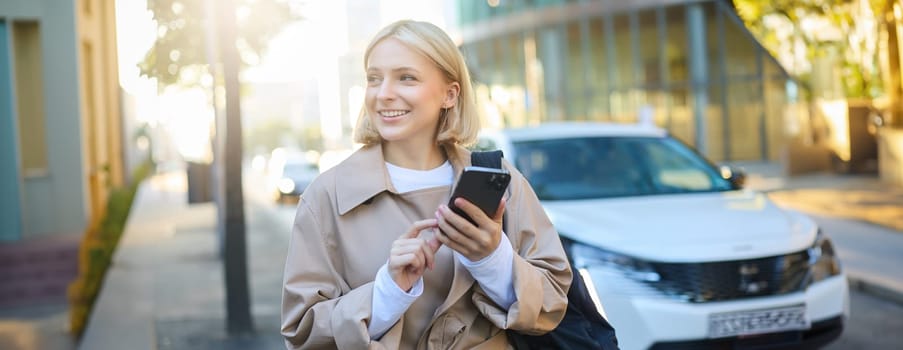Portrait of young woman using map application on smartphone, student with backpack, walks along street on sunny day, holds mobile phone in hand.