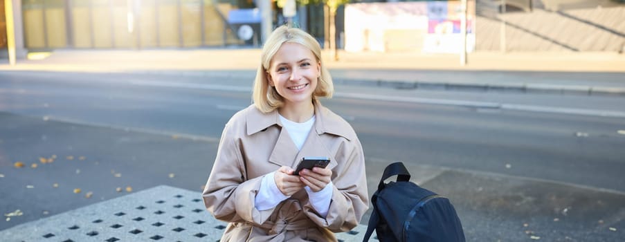 Portrait of smiling female model sitting on street bench with smartphone, waiting for friend, ordering a ride using mobile app.