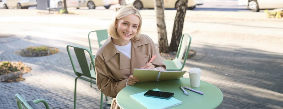 Portrait of beautiful blond woman, sitting in outdoor coffee shop, drawing in cafe in notebook, making sketches outside on street,