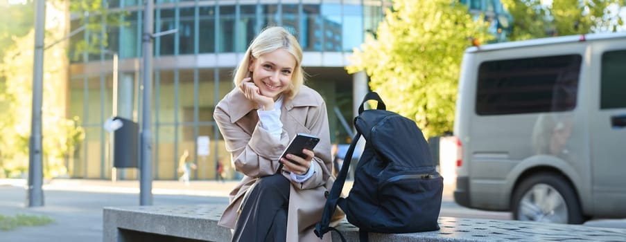 Lifestyle portrait of smiling female model sitting on street bench with backpack and mobile phone, waiting for friend and scrolling social media on smartphone.