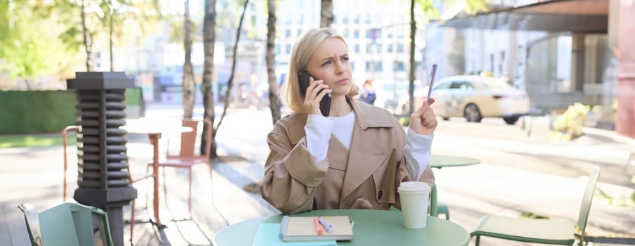Image of woman with confused, serious face, answers phone call, working in cafe outdoors, drinking coffee, frowning with pen in hand.