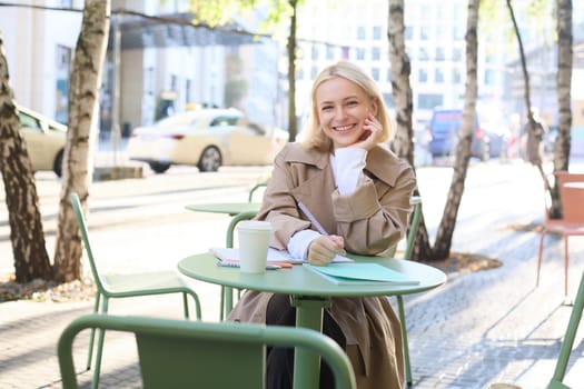 Portrait of young modern woman in stylish trench coat, sitting in outdoor cafe on sunny day, writing in her notebook, working or doing homework, drinking coffee.