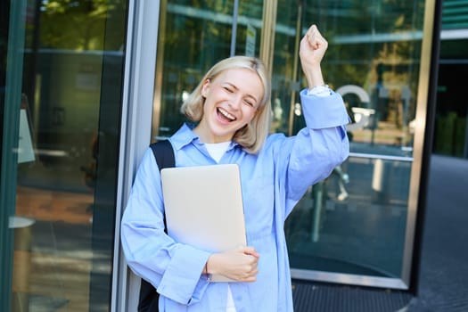 Carefree blond woman laughing, smiling and celebrating, posing with laptop near office, campus building, raising hand up in triumph, feeling excited. Copy space