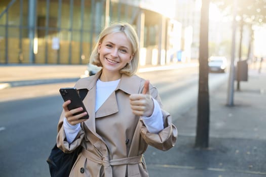 Portrait of young woman standing on street, gives her recommendations, shows thumbs up and using smartphone, smiling at camera.
