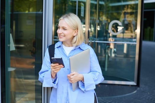 Image of stylish young woman, student walking out of the college campus, holding smartphone and laptop, has backpack on shoulder, waiting for someone outside with mobile phone.