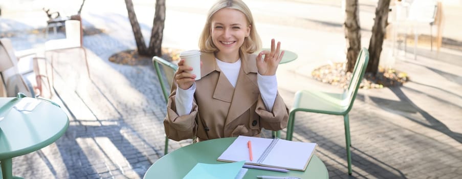 Cheerful young student, woman sitting in street cafe, holding cup of coffee, showing okay sign, ok gesture, doing homework, writing in notebook.