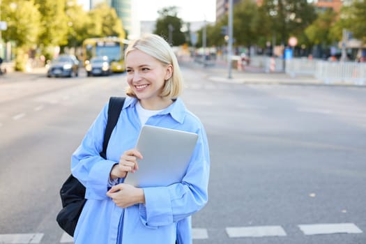 Beautiful female model in blue shirt, holding laptop computer and backpack, posing on street with skyscrapers and road behind, looking happy and confident.